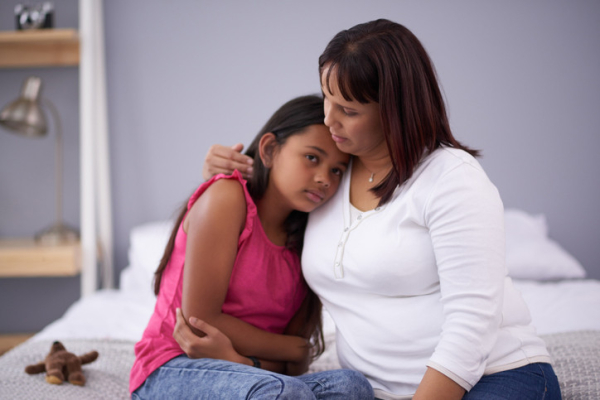 A child in a pink shirt and blue jeans with her arms around her stomach, sitting on a bed curled up against her mother; concept is stomach pain